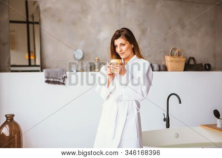 Beautiful Woman Relaxing In White Bathrobe Standing Near Bath With Cup Of Tea Or Coffee