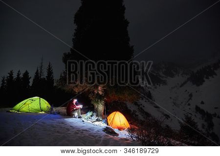 Man Sits Near A Tents At Night In The Winter In The Mountains