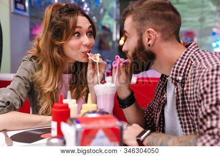 Photo of a cute positive young loving couple sit in retro bright cafe drinking cockail indoors.