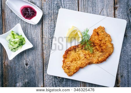 Traditional deep fried Wiener schnitzel from veal topside with cucumber salad and cranberry jam offered as top view on a modern design plate on a rustic wooden board 