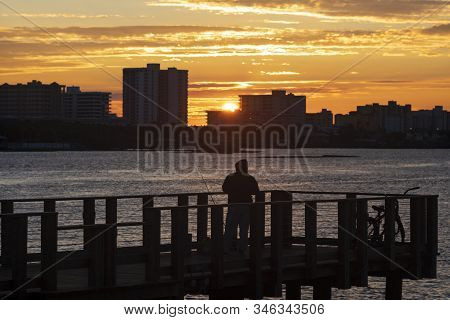 A Silhouetted Man Fishing Off A Pier In The Halifax River In Daytona Bearch, Florida As The Sun Rise