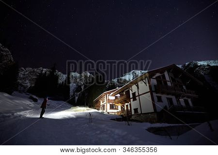 Man Tourist In The Mountains Shine To A White Chalet At Night Under A Sky Full Of Stars.