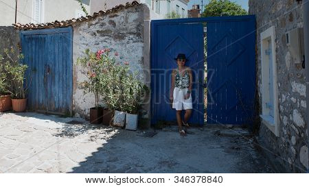 Asian Woman In Sunglasses And Hat Standing In Front Of Traditional Greek House With Blue Shutters An