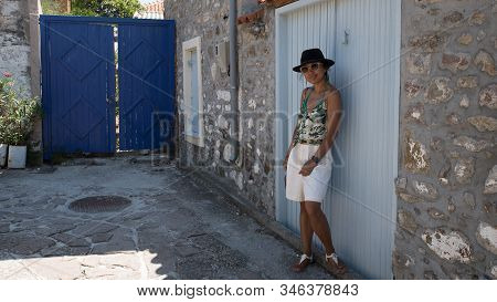Asian Woman In Sunglasses And Hat Standing In Front Of Traditional Greek House With Blue Shutters An