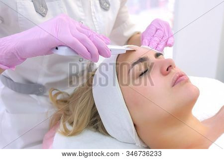 Cosmetologist Making Ultrasonic Face Cleaning Procedure For Young Woman In Clinic. Closeup Client Po