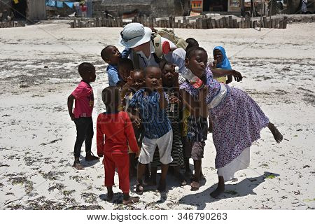 Pingwe, Zanzibar - October 6, 2019: European Man Tourist Surrounded Of Group The Funny African Child