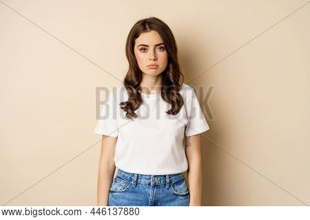 Sad And Tired Brunette Woman Looking Drained And Unamused, Standing With Pokerface Against Beige Bac