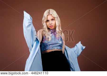 A Blonde In A Turquoise Jacket, In A Blue Jacket In Sequins, Poses In The Studio On A Brown Backgrou