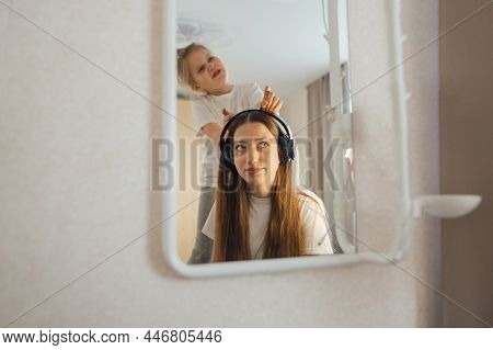Teenage Girl In Headphones In Front Of A Mirror, Little Sister Interferes, Grimaces.