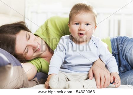 Adorable joyful baby boy sitting in bed next to his mother while she is embracing him lying down and looking at him.