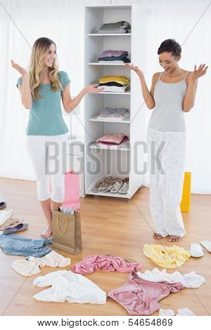 Two happy young women looking down at shopping bag and scatted clothes on floor at home
