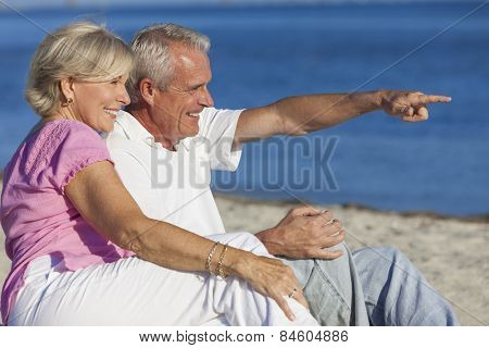 Senior man and woman couple sitting on a deserted beach pointing to the sea