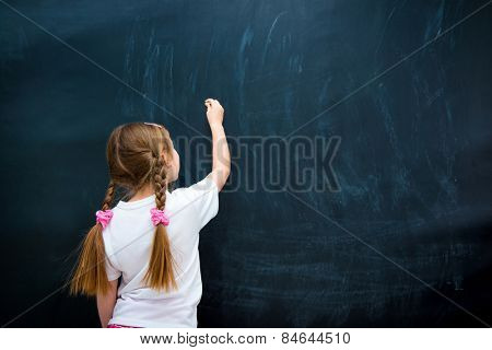little girl in a white T-shirt with pigtails writing with chalk on chalkboard