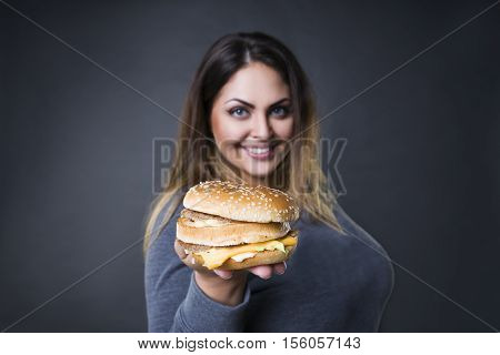 Happy beautiful young plus size model posing with hamburger on a gray studio background xxl woman eating burger fast food and unhealthy nutrition concept