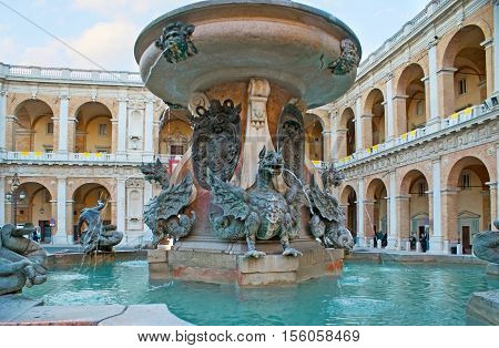 LORETO ITALY - OCTOBER 6 2012: The medieval stone fountain with bronze dragons tritons and dolphins in the Madonna Square with the Apostolic Palace on the background on October 6 in Loreto.