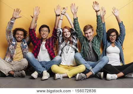 Multiethnic group of happy young people sitting together with raised hands over yellow background