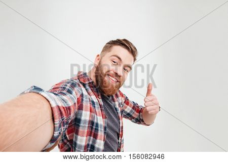 Close up portrait of a happy casual man taking selfie and showing thumbs up gesture over white background