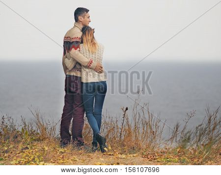 Young attractive couple in knitted sweaters standing on a cliff at sea shore and hugging