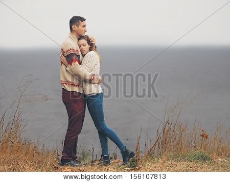Young attractive couple in knitted sweaters standing on a cliff at sea shore and hugging