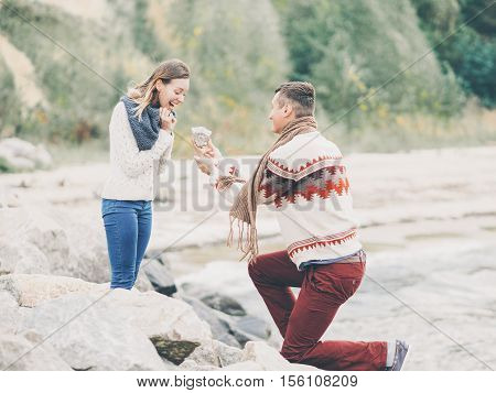 Young attractive couple in knitted sweaters on a cliff on sea shore man jokingly proposes and gives his girfriend a rock creative toning