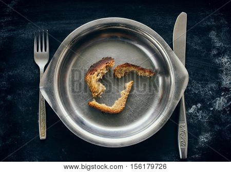 three crust of bread in metal plate fork and knife on black dirty surface concept starvation