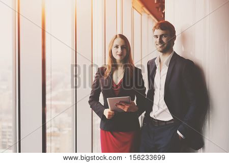 Group of business in office interior standing between window and column: smiling elegant man in formal suit and his female colleague in red dress and jacket holding tablet