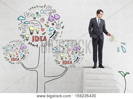 Businessman in glasses is watering a sketch of a plant while standing on stairs near concrete wall with business idea trea.