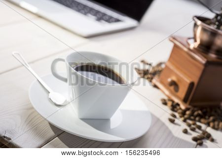 Close up of a cup of coffee with a saucer and a spoon. Coffee grinder and beans are lying near a laptop on wooden desk