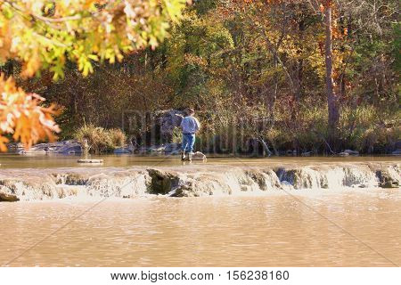 An older man, facing away, wearing blue jeans, blue plaid shirt and wading shoes, is standing in the Blue River of Oklahoma, fly fishing in fall. Trees with fall colors in background and autumn leaves and small waterfall is in foreground.