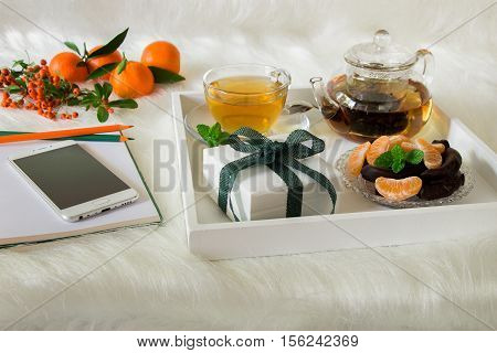 Green mint tea mandarin slices chocolate and gift box on tray near notebook pens mobile mandarins mountain ash sprig on white artificial fur background. Happy time break and gift.