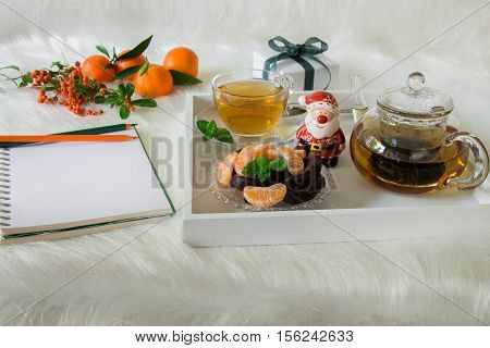 Green mint tea chocolate mandarin slices and Santa Claus on tray near notebook pencils mandarins mountain ash on white artificial fur background. Time break notebook surprise preparation.