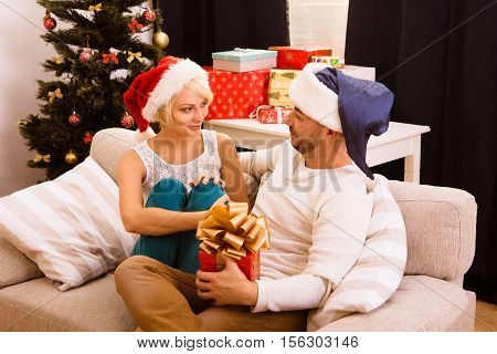 Picture of happy and romantic Christmas couple celebrating New Year while sitting on sofa or couch at home. Man looking at his wife and ready togive present or gift to her.