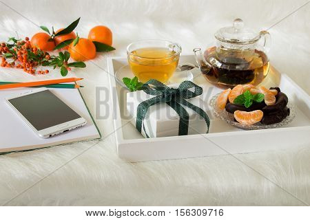 Green mint tea, mandarin slices, chocolate and gift box on tray, near notebook, pens, mobile, mandarins, mountain ash sprig on white artificial fur background. Happy time break and gift.