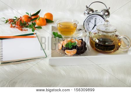 Green mint tea, chocolate, mandarin slices and alarm clock on tray near notebook, pencils, mandarins, mountain ash on white artificial fur background. Morning time breakfast notebook plan preparation.