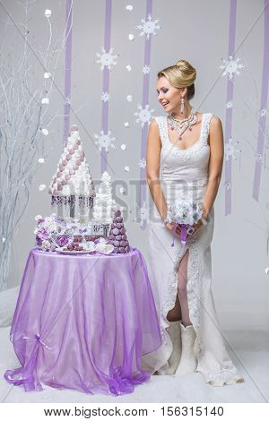 Beautiful young woman in wool wedding dress holding bridal bouquet standing near small table with big wedding croquembouche cake. Winter bride on snow flakes background. Copy space.
