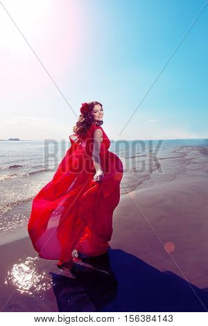 Beautiful girl in a bright red dress by the sea. Against the background of the summer lanshaft. Young romantic woman in the sunshine, sunset.