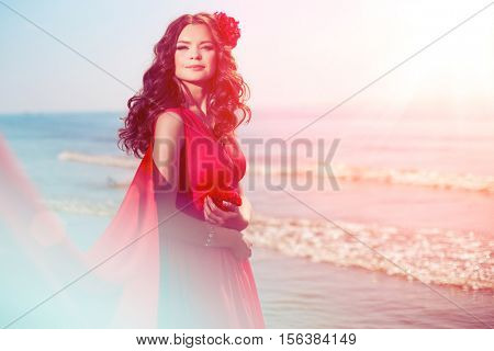 Beautiful girl in a bright red dress by the sea. Against the background of the summer lanshaft. Young romantic woman in the sunshine, sunset.
