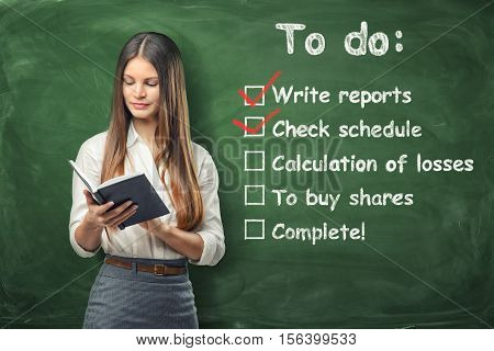 A young woman holding her day planner standing near the list of things to do at work written on the green chalkboard. Daily routine and time-management. Check schedule and write reports. To allocate resources appropriately.