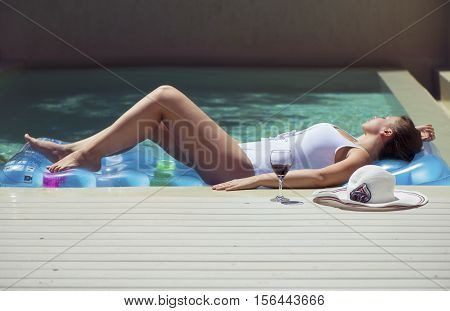 female in a swimsuit lying near the pool with hat and wine