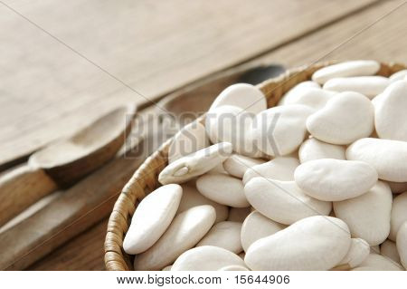 Basket full of white bean. Wooden cutlery next to it. All on wooden table.