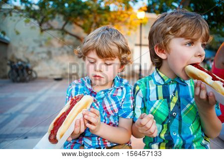 Two little kid boys eating hot dogs outdoors. Siblings enjoying their meal. Hotdog as nhealthy food for children.