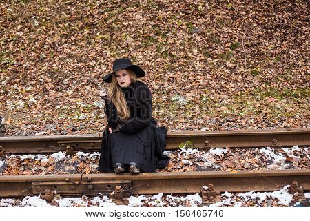 Young woman in a long black dress and hat smoking a cigarette while sitting on railroad tracks waiting for a train. The concept of loneliness and expectations. Travel alone.