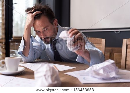 Need new ideas. Exhausted cheerless depressed man holding his head and crumpling a sheet of paper while dealing with a problem