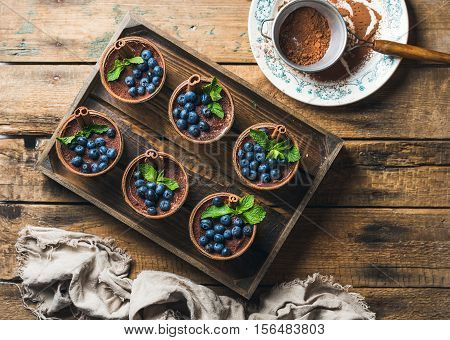 Homemade Tiramisu dessert in glasses with cinnamon, mint and fresh blueberries in wooden tray and sieve with cocoa powder in white plate over rustic wooden background, top view, copy space