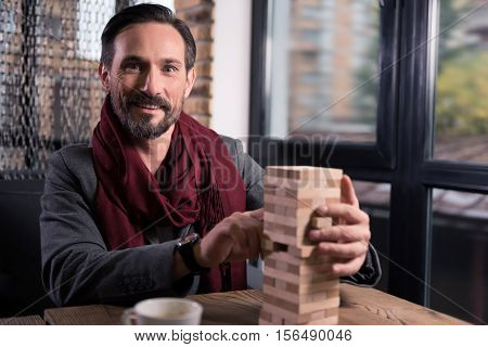 Pleasant entertainment. Happy handsome good looking man holding a wooden tower and smiling while playing jenga