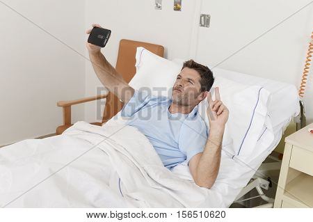 young attractive man lying on bed hospital clinic holding mobile phone taking self portrait selfie photo in optimist recovery concept and health care positive image