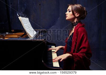 Side mid shot of charming woman in eveningwear with lace black necklace and red scarf playing solo without any accompaniment. Sitting on chair at the piano