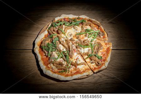 Pork pizza with mushrooms and cheese decorated with leaves of arugula to cut off a piece served on a wooden tray on old wood table of In the dark
