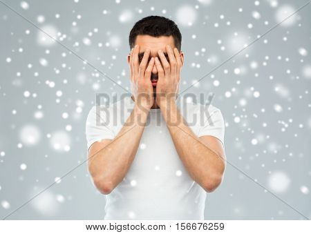 people, fear, emotions, winter and stress concept - man in white t-shirt covering his face with hands over snow on gray background