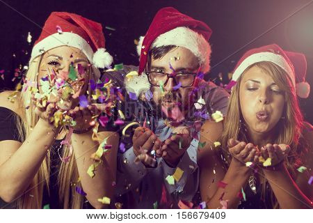 Three young people wearing Santa's hats blowing colorful conffeti at midnight at New Year's Eve party